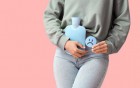 Person hält eine Wärmflasche auf den Unterbauch und ein trauriges Symbol in der Hand vor rosa Hintergrund.