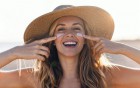 Lächelnde Frau mit Sonnenhut trägt Sonnencreme unter den Augen auf.